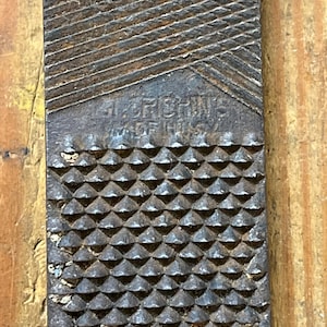 May include: Close-up of a well-used, rectangular rasp file. The file has a textured surface with diagonal and diamond-shaped patterns. The text "J.F. CRISHON'S MADE IN USA" is visible. The file is a tool for shaping wood or metal.
