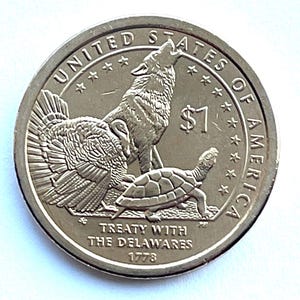 May include: A silver dollar coin featuring a howling wolf, a turkey, and a turtle. The coin is inscribed with "United States of America" and "Treaty with the Delawares 1778". The denomination is $1.