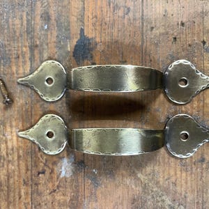 May include: Two antique brass drawer pulls with decorative end pieces and a textured center band. The pulls are accompanied by several screws, all resting on a weathered wooden surface. Hardware for furniture.