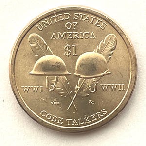 May include: A gold-colored $1 coin featuring two World War I helmets, crossed feathers, and the text "UNITED STATES OF AMERICA". The coin also includes the text "WWI", "WWII", and "CODE TALKERS".
