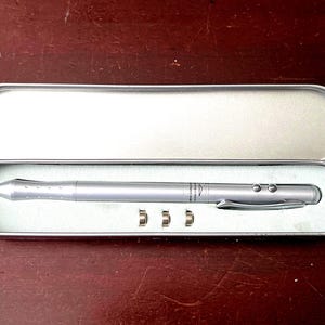 May include: A silver laser pointer pen with three small silver attachments, presented in an open, rectangular, silver metal case. The pen has a clip and two buttons. The case is on a dark brown surface.