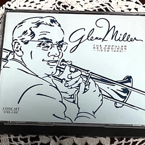 May include: A CD set featuring a light blue illustration of Glenn Miller playing the trombone. The text reads "Glenn Miller" and "The Popular Recordings (1938-1942)". The CD case is a 3-disc set.