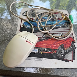 May include: A Compaq computer mouse with a cream-colored body and a red Compaq logo. The mouse is connected to a cream-colored cable and rests on a mousepad featuring illustrations of classic cars in red and blue.