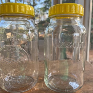 May include: Two clear glass Mason jars with yellow lids. One jar is embossed with "golden harvest" and a circular design, the other with "MASON". The jars are empty and sit on a wooden surface.