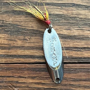 May include: A vintage fishing lure with a silver-colored, teardrop-shaped body. The lure is marked "MR CHAMP" and has a red and yellow feather tail. The lure is designed for catching fish.