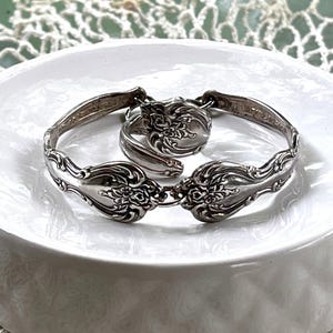 May include: A silver-toned bracelet and ring set, crafted from repurposed silverware. The bracelet features ornate floral detailing on the spoon bowls, connected by hinges. The ring mirrors the design, creating a cohesive vintage jewelry set.