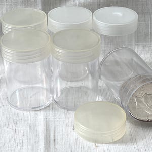 May include: Six clear plastic containers with white lids. The containers are stacked on top of each other. One container has a silver coin inside.