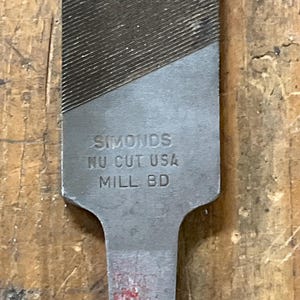 May include: Close-up of a metal file with the text "SIMONDS NU CUT USA MILL BD" stamped on the handle. The file has a textured surface with diagonal grooves. The handle has a rectangular shape.