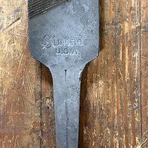 May include: Close-up of a vintage metal file with the text "Simonds U.S.A." stamped on the handle. The file has a textured surface on the top portion and a long, narrow handle. The background is a wooden surface.