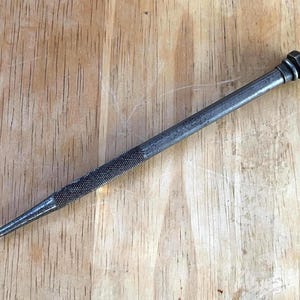 May include: A metal pin punch tool with a textured grip and a pointed tip. The tool is silver and has a hexagonal head. The tool is lying on a wooden surface.