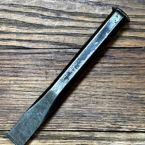 May include: A black metal chisel with a rectangular blade and a beveled edge. The chisel has a long, slightly tapered body and a flat striking end. The tool shows signs of wear, with some areas of the black finish worn away.