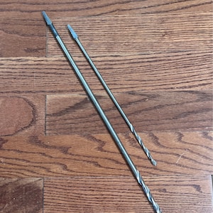 May include: Two silver drill bits of different sizes are laid diagonally across a wooden surface. The larger bit has a spiral design along its length, while the smaller one is straight. Both have pointed tips, designed for drilling into materials.