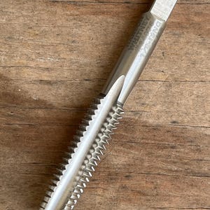 May include: A silver-colored tap tool with a ridged cutting edge and a hexagonal head. The tool has the text "WHITNEY METAL TOOL" and "DNG 1/2-13" etched on its surface. The tool is designed for threading and is set against a wooden background.