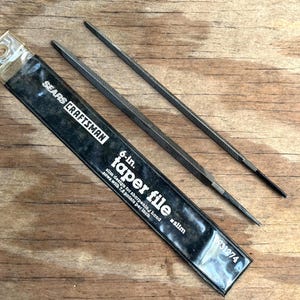May include: Two metal taper files, one in a clear plastic sleeve, are displayed on a wooden surface. The sleeve is labeled "SEARS CRAFTSMAN" and "6-in. taper file." The files are slim and designed for sharpening hand saws.