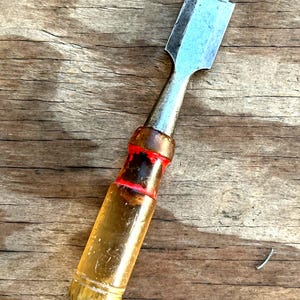 May include: A woodworking chisel with a metal blade and a clear, yellow handle. The handle has a red band near the blade. The chisel is resting on a weathered wooden surface.