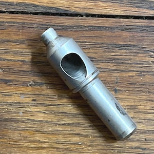 May include: A silver-colored metal tool with a cylindrical body and a large, oval-shaped opening. The tool has a slightly tapered end and appears to be used for creating holes or shaping materials. The tool is resting on a wooden surface.