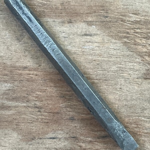 May include: A dark gray metal chisel with a faceted body and a flat, beveled cutting edge. The tool is designed for woodworking or metalworking. The chisel is shown against a wooden background.