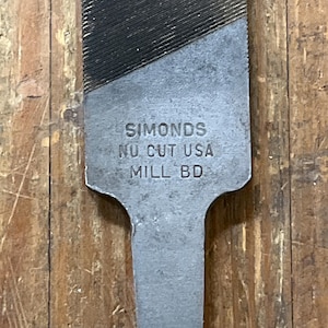 May include: Close-up of a metal file with the text "SIMONDS NU CUT USA MILL BD" etched on the surface. The file has a textured, ridged cutting surface and a flat, elongated handle. The tool is designed for shaping and smoothing materials.