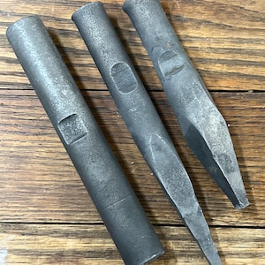 May include: Three dark gray metal chisels with cylindrical handles and angled cutting edges. The tools are arranged diagonally on a wooden surface, showcasing their industrial design and craftsmanship. The chisels appear to be well-used.