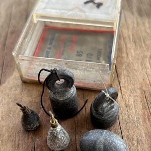 May include: A collection of vintage fishing weights in various shapes and sizes, including teardrop, cylindrical, and oval forms. The weights are dark gray and black, with some featuring metal attachments. A clear plastic box is open in the background.