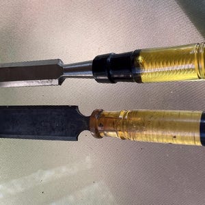 May include: Two wood chisels with metal blades and yellow-tinted, cylindrical handles. One chisel has a black handle end, and the other has a brown handle end. The blades are dark gray and rectangular.