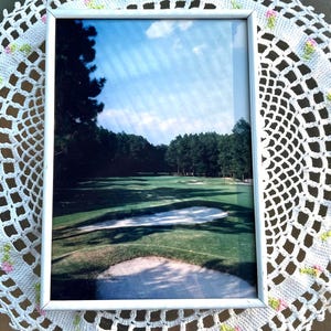 May include: A white framed photograph of a golf course with a sand trap in the foreground. The photo is displayed on a white crocheted doily with pink and green flowers.