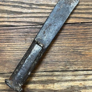 May include: A well-worn, antique metal chisel with a rectangular blade and a cylindrical handle. The tool shows signs of age and use, with a weathered appearance and a dark, metallic color. The chisel is positioned diagonally on a wooden surface.