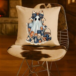 Puede incluir: Un cojín cuadrado de color beige con un gráfico central de un fantasma que lleva un lazo grande estampado y una capa estilo patchwork en tonos azules y naranjas. El fantasma sostiene un pequeño cubo de calabaza naranja para dulces.