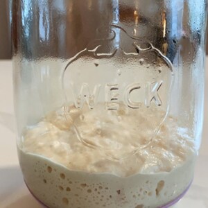 May include: A glass jar with a rubber band around the top, filled with a white, bubbly substance. The jar has the word "WECK" embossed on the side.