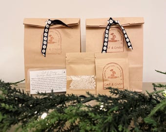 May include: Two brown paper bags with daisy-patterned ribbon and logo stickers, alongside smaller packets of oats. The bags have the text "OAT & DAISY" on them. A sprig of green garland is in the foreground.