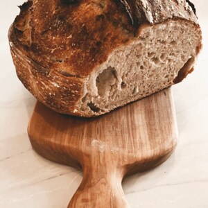 May include: A freshly baked loaf of sourdough bread with a crispy crust and a soft, airy crumb. The bread is sitting on a wooden cutting board with a handle.