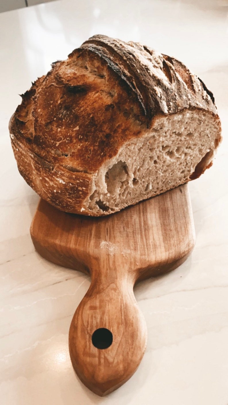May include: A freshly baked loaf of sourdough bread with a crispy crust and a soft, airy crumb. The bread is sitting on a wooden cutting board with a handle.