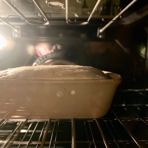 May include: A loaf of bread baking in a white ceramic loaf pan inside an oven with a rack.