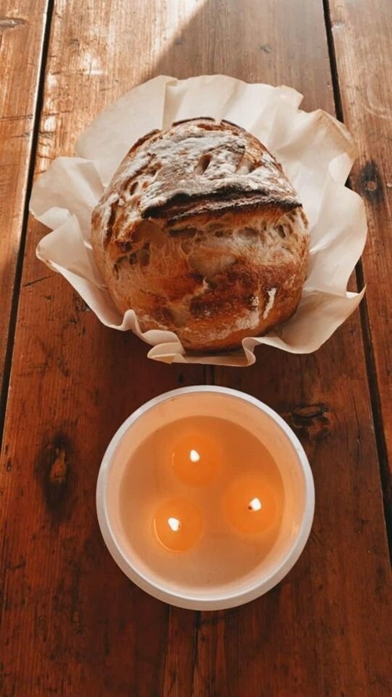 May include: A freshly baked loaf of bread sits on a white paper doily on a wooden table. A white candle holder with three lit candles is in the foreground.