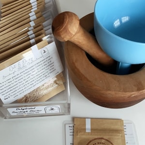 May include: A close-up of a brown paper packet labeled "Dehydrated Sourdough Starter" with the text "Oat & Daisy" and a mushroom graphic. The packet is next to a wooden mortar and pestle with a light blue ceramic bowl.