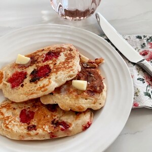 May include: Three golden brown pancakes with red fruit filling and a pat of butter on top, served on a white plate with a glass of water and a silver knife on a white surface.