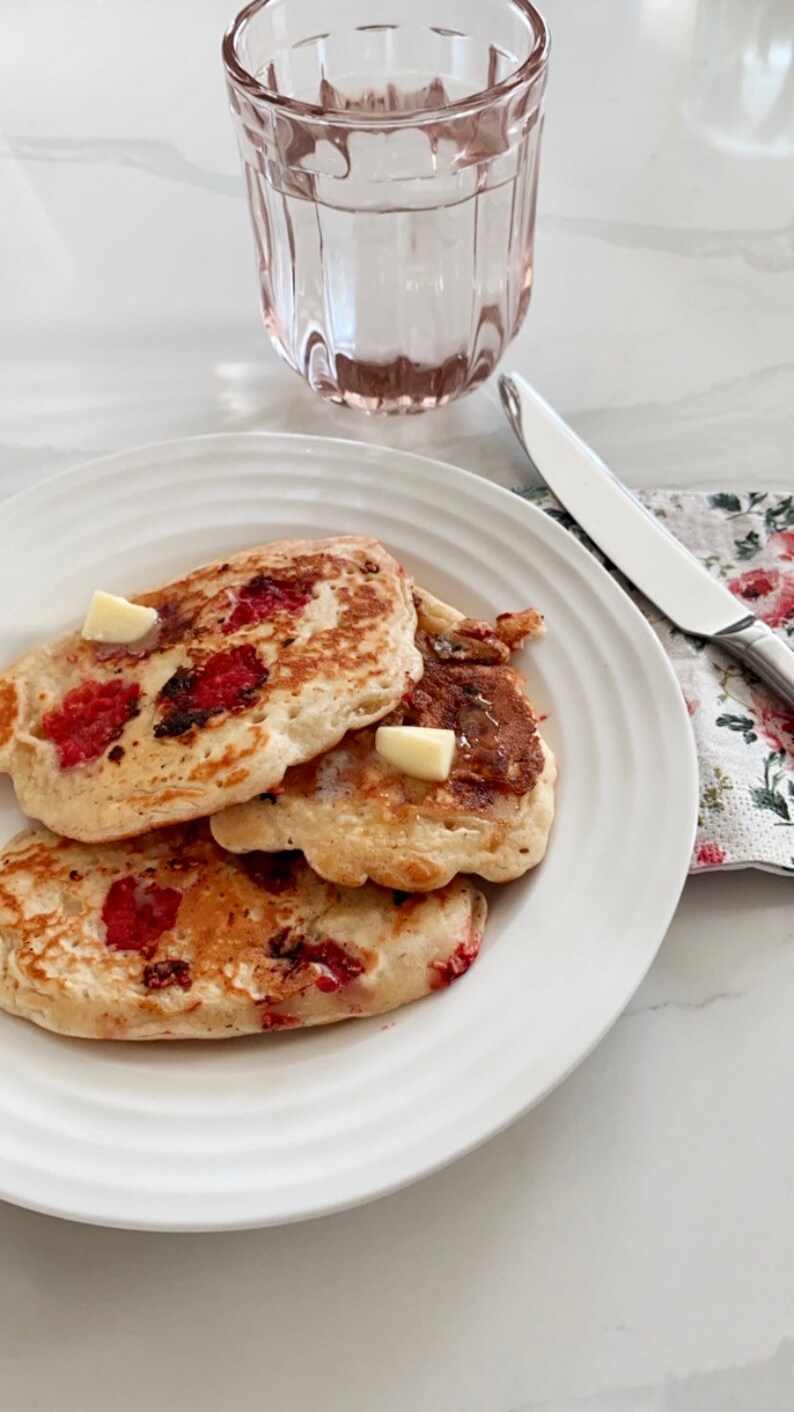 May include: Three golden brown pancakes with red fruit filling and a pat of butter on top, served on a white plate with a glass of water and a silver knife on a white surface.