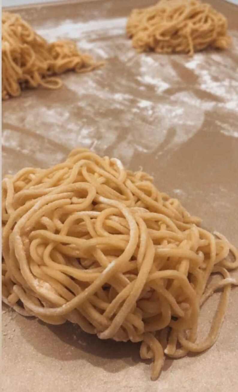 May include: A close-up of three piles of fresh, golden-brown noodles on a brown surface. The noodles are arranged in a loose, tangled pile, and they appear to be dusted with flour.