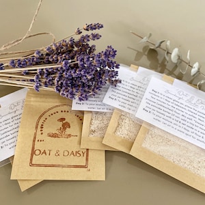 Peut inclure: Deux petits sachets en papier brun contenant de la poudre blanche. Les sachets sont étiquetés "Oat & Daisy Sourdough Starter" et contiennent des instructions pour faire un levain. Les sachets sont posés sur une surface brun clair avec un bouquet de fleurs de lavande séchées.