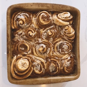 May include: A close-up of a baking dish filled with freshly baked cinnamon rolls, glazed with a sweet icing. The rolls are golden brown and have a swirl pattern.