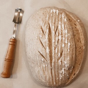 May include: A close-up of a loaf of bread with a tree-shaped scoring pattern on top. A bread lame, a tool used for scoring bread, is also in the image.