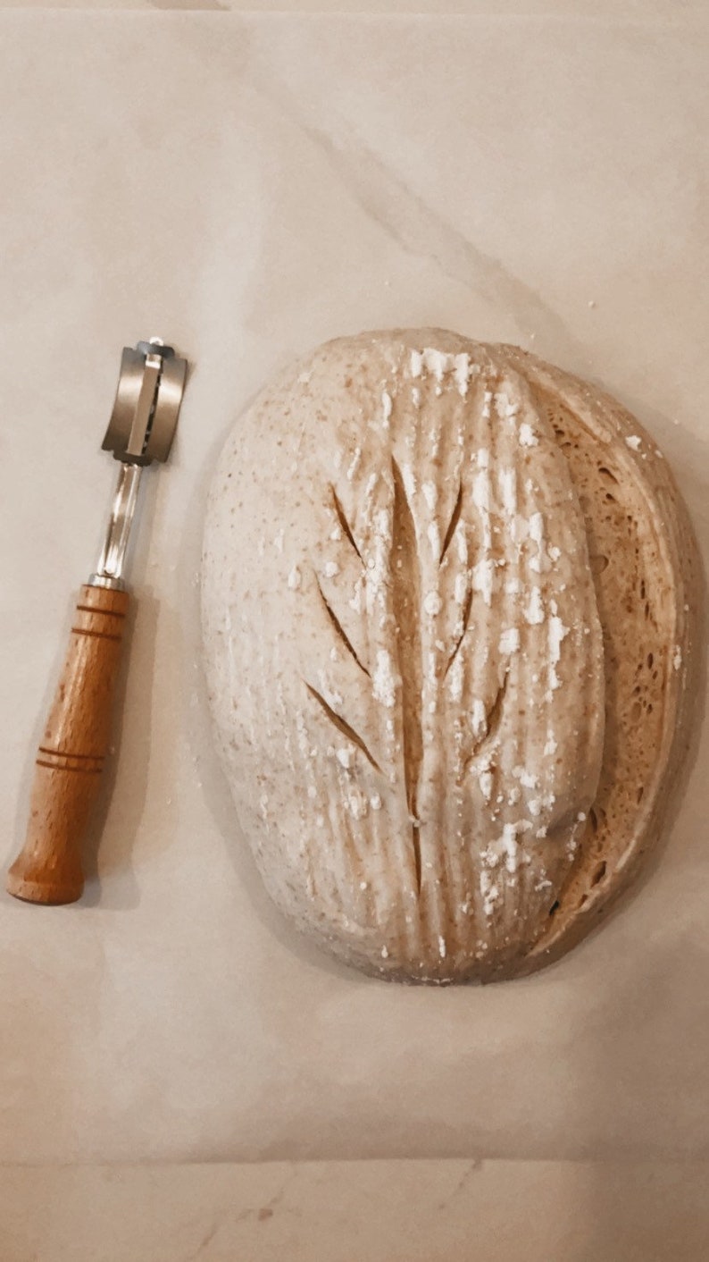 May include: A close-up of a loaf of bread with a tree-shaped scoring pattern on top. A bread lame, a tool used for scoring bread, is also in the image.
