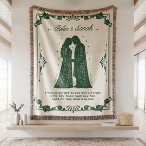 May include: A cream-colored woven throw blanket featuring a dark green decorative border and a central illustration of two figures in robes. The text reads John & Sarah 20 26 and I would rather share one lifetime with you than face all the ages of this world alone.