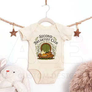 Puede incluir: Un body de bebé de manga corta color crema con un estampado gráfico que incluye el texto "SECOND BREAKFAST CLUB" sobre una puerta redonda verde, una mesa de madera con una tetera, una taza y dos cruasanes, rodeado de ramas verdes.