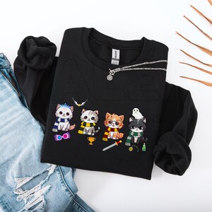 Puede incluir: Sudadera negra con un gráfico de cinco gatos de dibujos animados vestidos como personajes de la serie Harry Potter. Los gatos llevan bufandas y sombreros, y uno de ellos sostiene una varita mágica. El texto "Hufflepuff" es visible en la sudadera.
