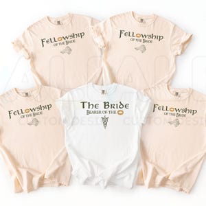 May include: Five short-sleeved t-shirts in cream and white. Four cream shirts feature the text Fellowship of the Bride with a ring and leaf graphic. One white shirt features the text The Bride Bearer of the Ring with a leaf emblem graphic.