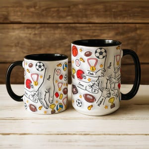 May include: Two white ceramic mugs with black handles and rims. The mugs are decorated with sports-themed illustrations, including footballs, soccer balls, and medals. The mugs have the names "Papa", "Noah", "Emma", "Matthew", and "Sarah" on them.