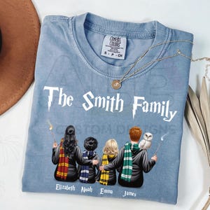 May include: A light blue t-shirt featuring the white text The Smith Family and a graphic of four individuals from behind, each wearing a different colored scarf and holding a wand. Below each figure are the names Elizabeth, Noah, Emma, and James.