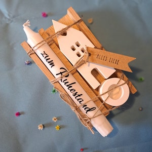 Könnte beinhalten: Ein weißes Keramikhaus mit einer Kerze und einem Holzsockel. Die Kerze ist mit Schnur umwickelt und trägt den Schriftzug "zum Ruhestand". Ein braunes Schild mit dem Text "Alles Liebe" ist am Haus befestigt.