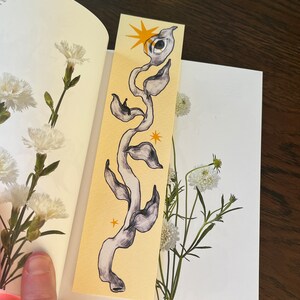 May include: A yellow bookmark with a black and white illustration of a vine with an eye and stars. The vine is stylized and has a whimsical feel.